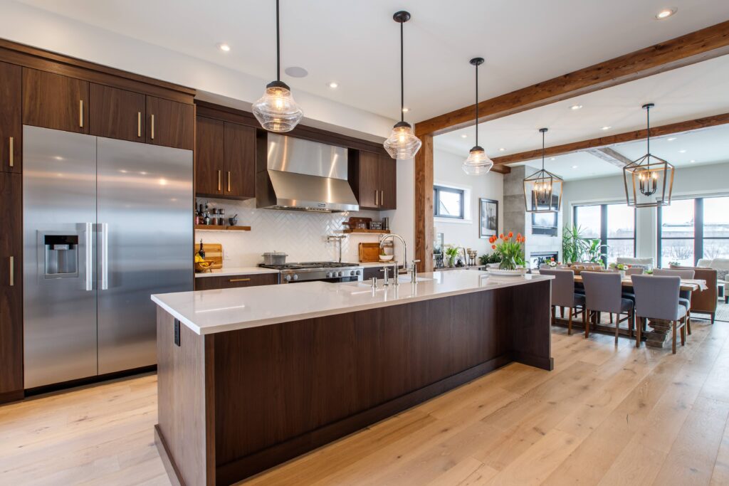 Modern kitchen with dark wood cabinets, a large stainless steel fridge, center island with white countertop, three pendant lights, light wood floor, and an open dining area with a table and chairs in the background.