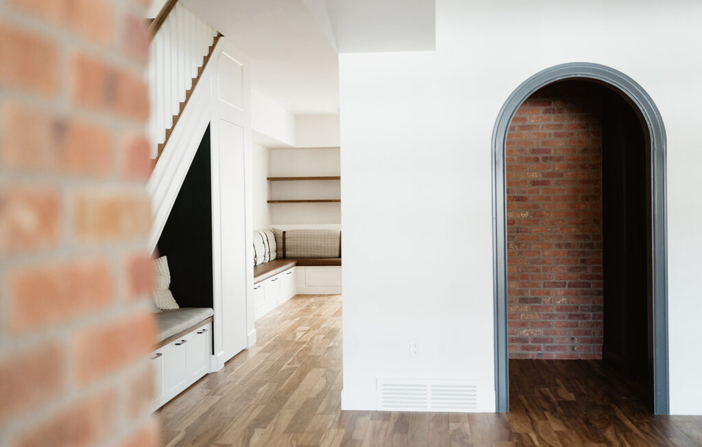 A bright interior features light wood floors, white walls, and built-in seating under a staircase. An arched doorway with a dark frame leads to a room with exposed red brick walls.