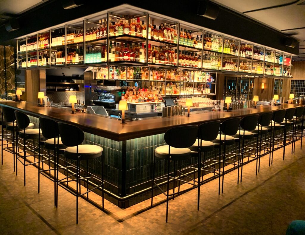 A modern, well-lit bar with high black chairs, a wooden counter, and shelves filled with liquor bottles overhead. Small lamps illuminate the bar, which is empty of people.