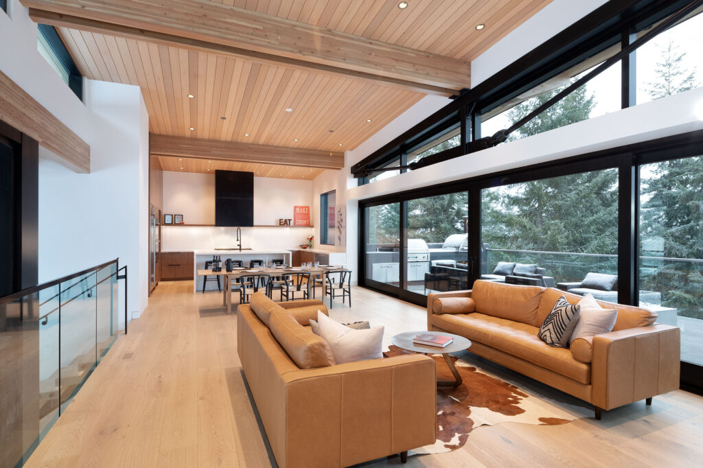 Modern open-concept living room and kitchen with tan leather sofas, large windows, wood ceilings, and a dining table. Glass doors open to a patio with trees visible outside. Neutral tones and natural light fill the space.