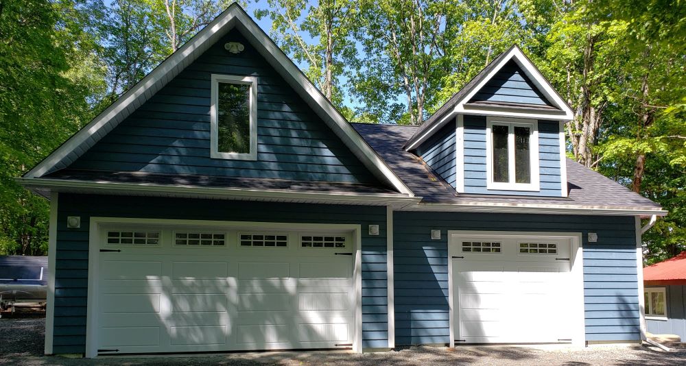 A two-car garage with blue siding, white trim, and two white garage doors is surrounded by trees. The garage has a steeply pitched roof and two upper windows, one above each door, with sunlight filtering through the leaves.