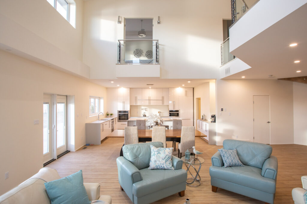 Bright, modern open-plan living space with light blue armchairs, a round glass table, dining area, and white kitchen. High ceilings, large windows, and wooden floors create an airy, spacious atmosphere.
