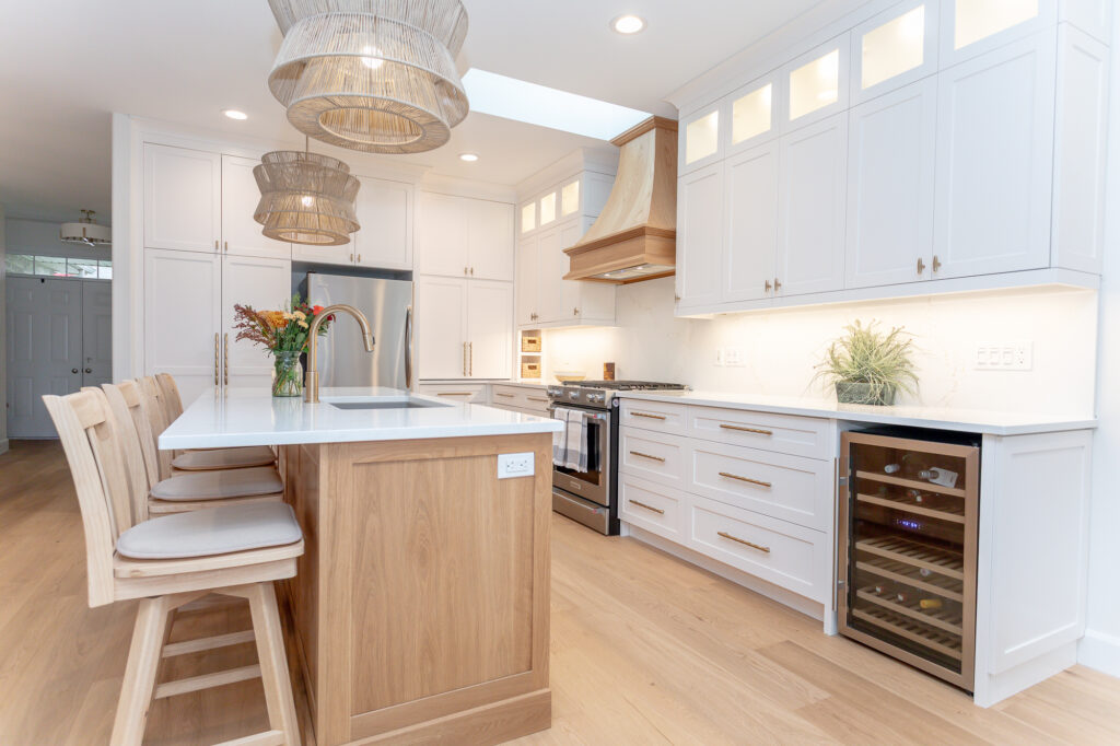 Modern kitchen with white cabinets, stainless steel appliances, a wooden island with three cushioned chairs, pendant lights, a built-in wine fridge, and light wood flooring. Flowers and a plant add decoration.