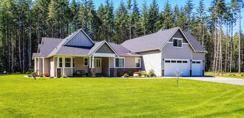 A modern, light-colored house with a front porch, large windows, and a two-car garage sits on a well-manicured lawn, surrounded by tall pine trees under a clear blue sky.
