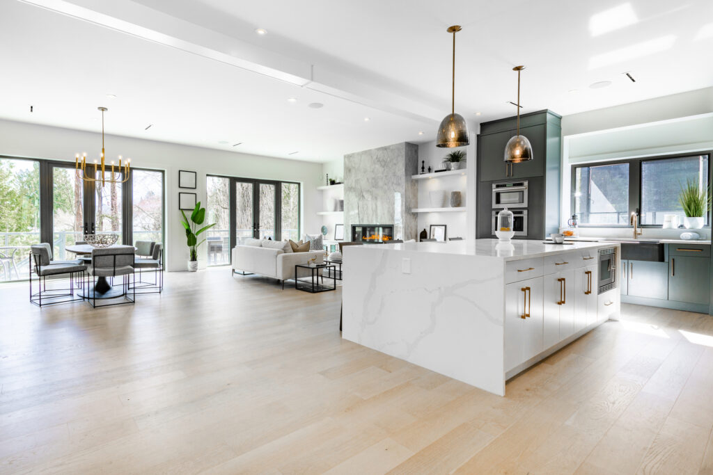 Modern open-concept kitchen and living area with white marble island, pendant lights, light wood floors, large windows, gray cabinets, and a dining area with a chandelier and outdoor view.