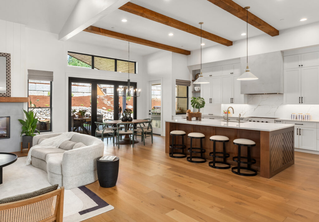 Modern open-concept kitchen and living area with wood beams, white cabinets, marble countertops, barstools, a dining table near large windows, and light wood flooring.