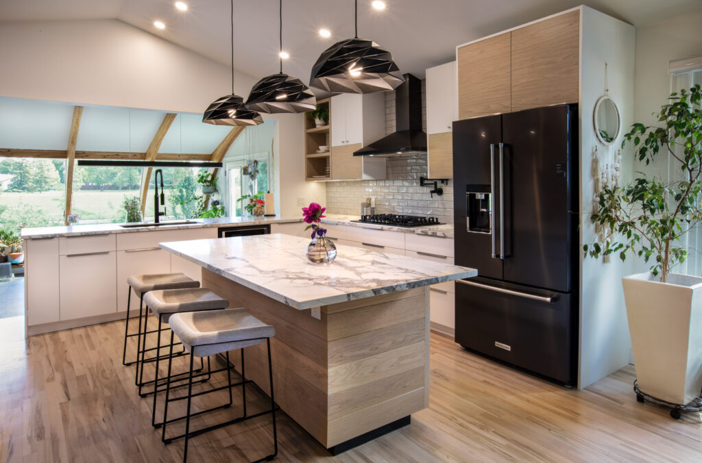 Modern kitchen with white cabinets, a marble island with stools, black pendant lights, a black refrigerator, gas stove, wooden accents, large windows, plants, and natural light. A vase with flowers sits on the island.