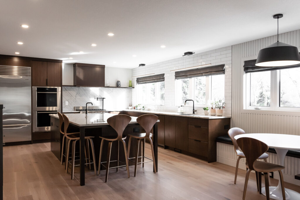 Modern kitchen with wood cabinets, a large island with four wooden chairs, stainless steel appliances, and big windows letting in natural light. There’s a dining table with chairs and potted plants by the windows.