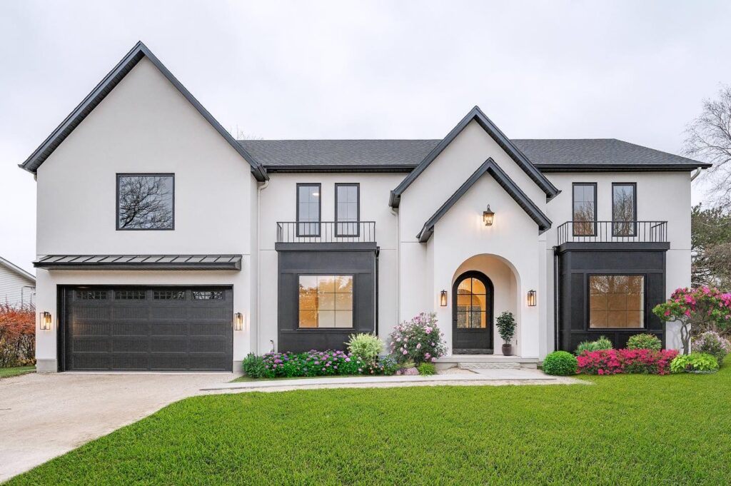 Modern two-story house with white exterior, black trim, double front doors, and a black garage. Flowering bushes and manicured green lawn decorate the front yard under a cloudy sky.