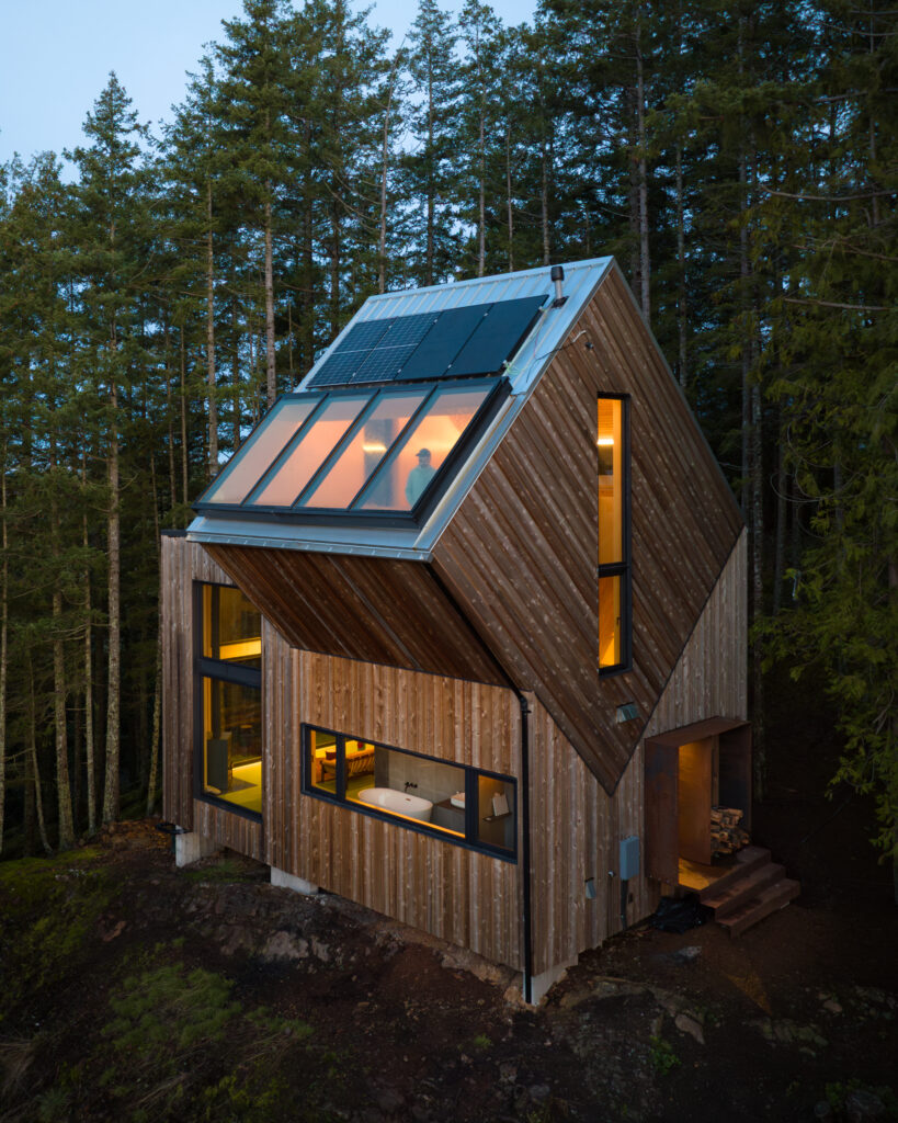 A modern wooden cabin with large windows and rooftop solar panels sits on a forested slope at dusk, glowing warmly from interior lights and reflecting the sky in the upper windows.