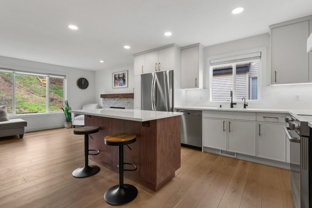 Modern kitchen with white cabinets, stainless steel appliances, wood flooring, a center island with two black stools, and large windows letting in natural light. Living area with a sofa and plant visible in the background.