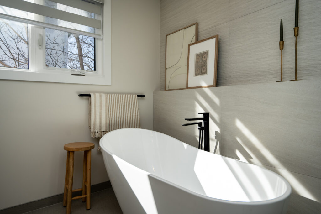 Modern bathroom with a white freestanding tub, black faucet, wooden stool, beige towel on a black rack, and framed art on a tiled wall. Sunlight streams in through a window with sheer blinds.