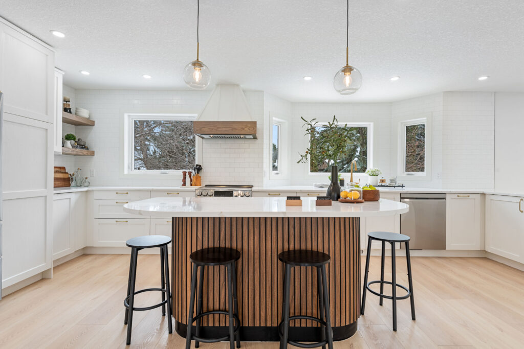 Modern kitchen with white cabinets, wood accents, a large island with striped wood paneling, three black stools, pendant lights, and decor including a plant and fruit on the counter. Large windows let in natural light.