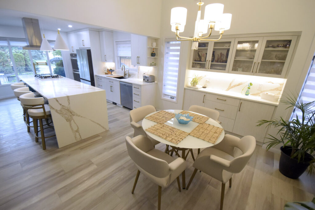 Modern kitchen and dining area with white cabinets, marble countertops, a large island, and beige upholstered chairs around a small round table. Pendant lights and a chandelier provide lighting; plants and decor add a cozy touch.