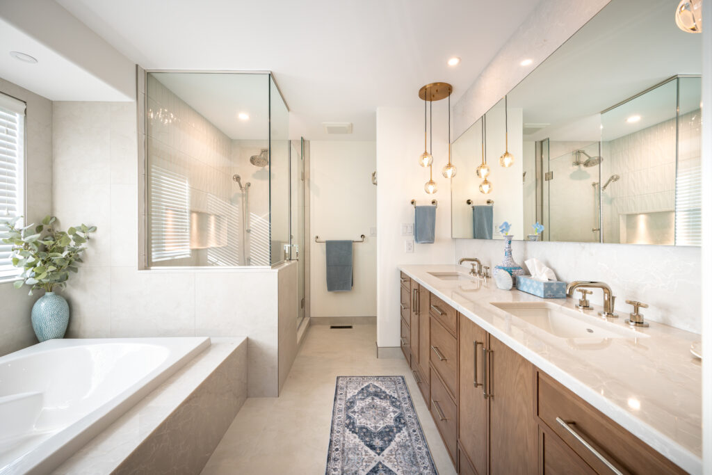 Modern bathroom with a glass-enclosed shower, double sinks with wooden cabinets, a bathtub near a window with blinds, hanging pendant lights, and decorative vases on the counter and tub ledge.