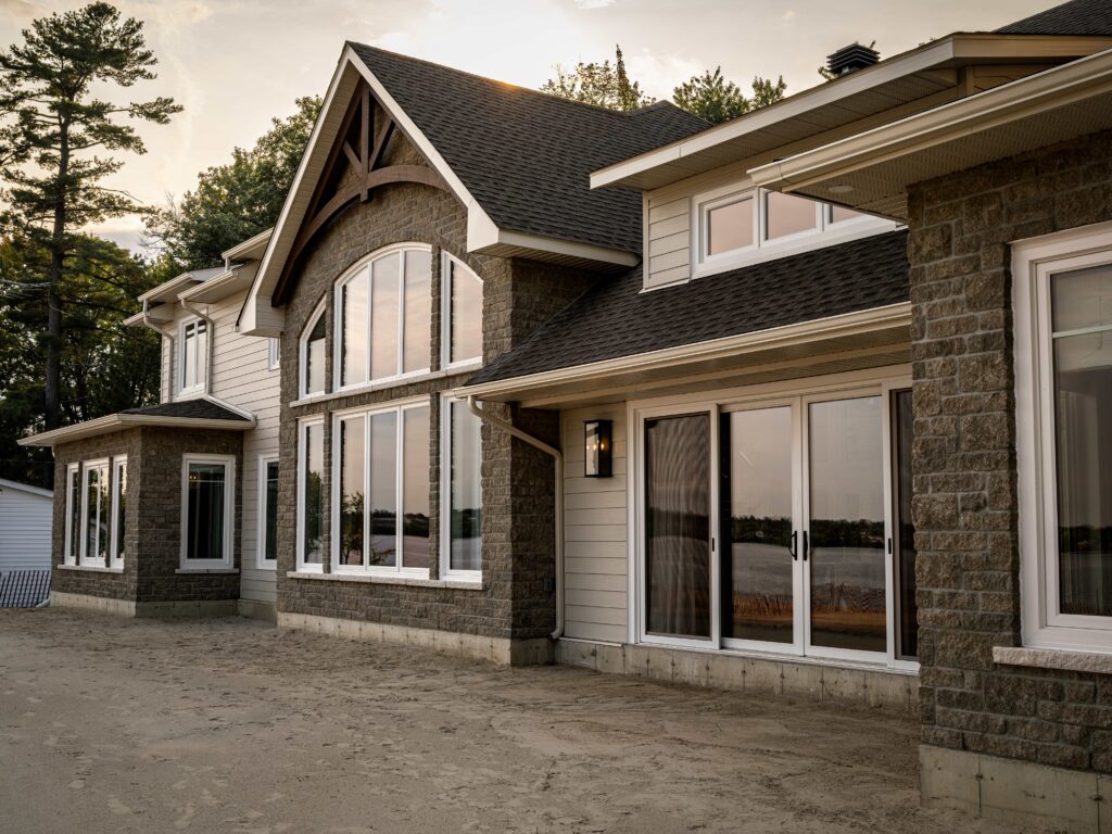 A modern house with large floor-to-ceiling windows, stone and siding exterior, and sliding glass doors, reflecting the sunset and trees. The house sits on a sandy lot near a body of water.