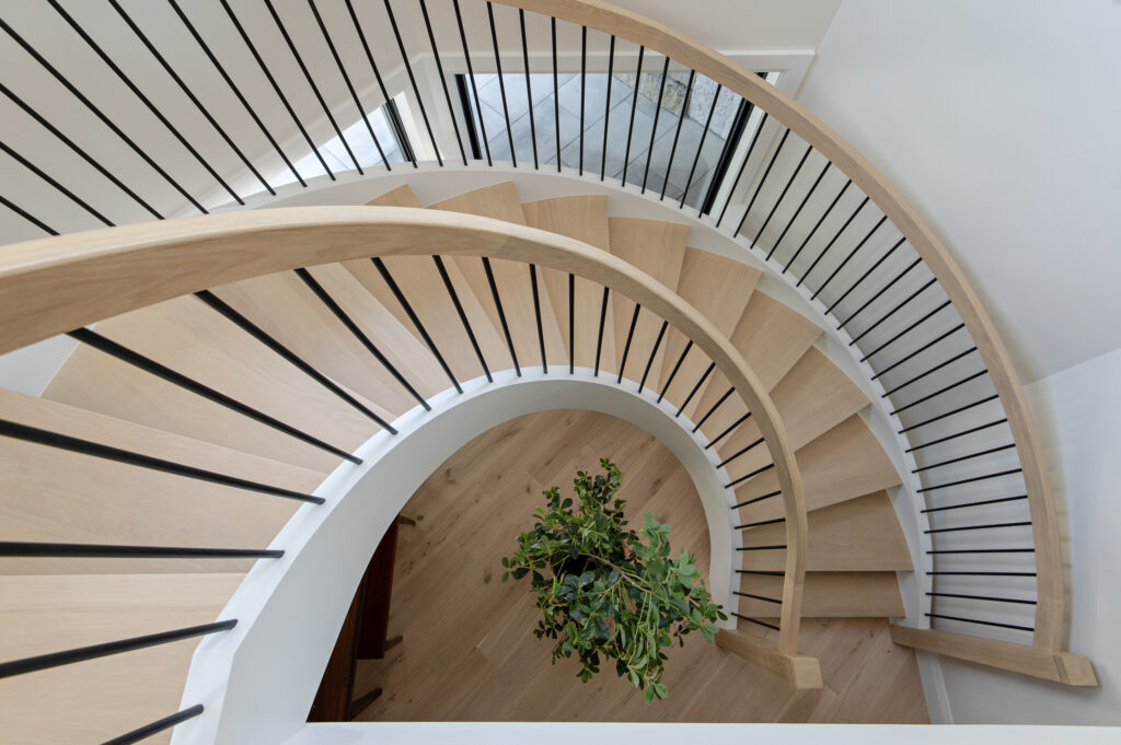 A top-down view of a modern spiral staircase with light wood steps and black vertical railings, curving around a green potted plant on a light wood floor below.