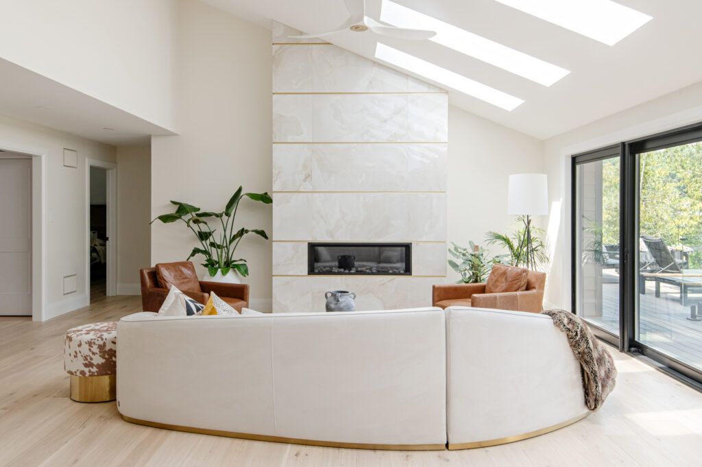 A modern, bright living room with a curved white sofa, two brown armchairs, a sleek marble fireplace, large windows, indoor plants, and light wood flooring under skylights.