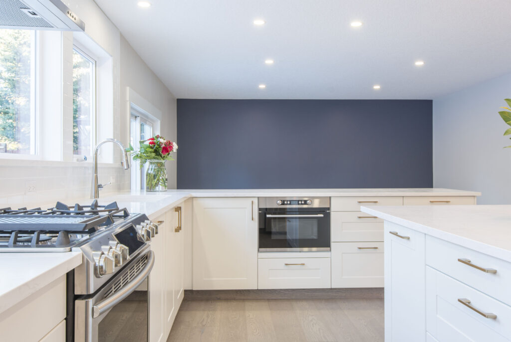 Modern kitchen with white cabinets, stainless steel stove, built-in oven, and a vase of flowers by a large window. The room has bright lighting and a dark accent wall in the background.