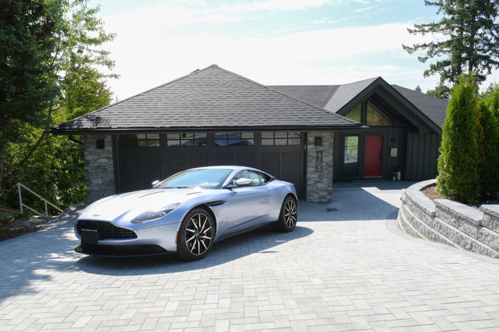 A silver sports car is parked on a light gray brick driveway in front of a modern house with stone and dark siding, surrounded by trees and greenery under a partly cloudy sky.