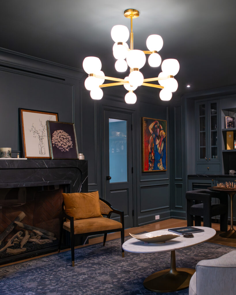 A modern living room with dark walls, a gold and white chandelier, a tan chair, a white oval coffee table, a fireplace with artwork on the mantel, and colorful paintings on the wall.