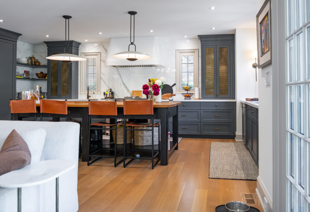 Modern kitchen with gray cabinets, a large island with four leather barstools, pendant lights, fresh flowers, wooden floors, and marble backsplash. Natural light streams in through a window above the sink.
