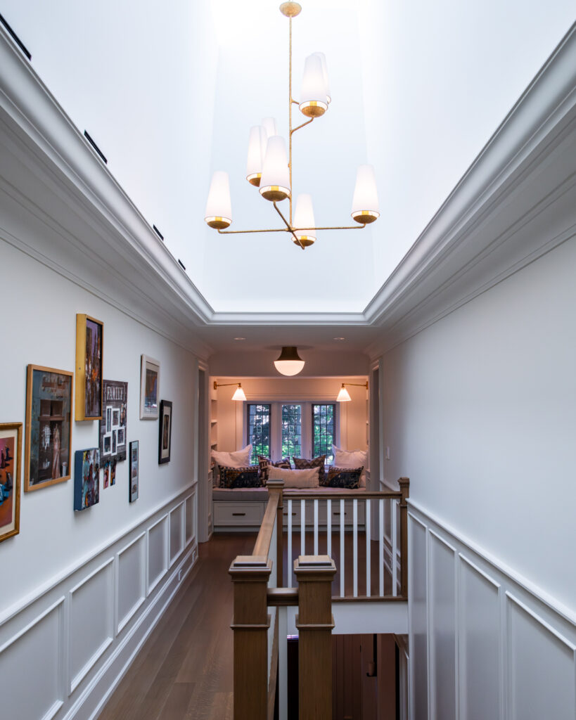A bright hallway with white walls and wainscoting features family photos on the left, a modern chandelier above, and a cozy reading nook with cushions in front of windows at the end.
