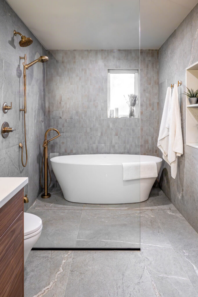 Modern bathroom with a freestanding white bathtub, gold fixtures, gray tiled walls and floor, a small window above the tub, white towels on a rack, and potted plants for decor.