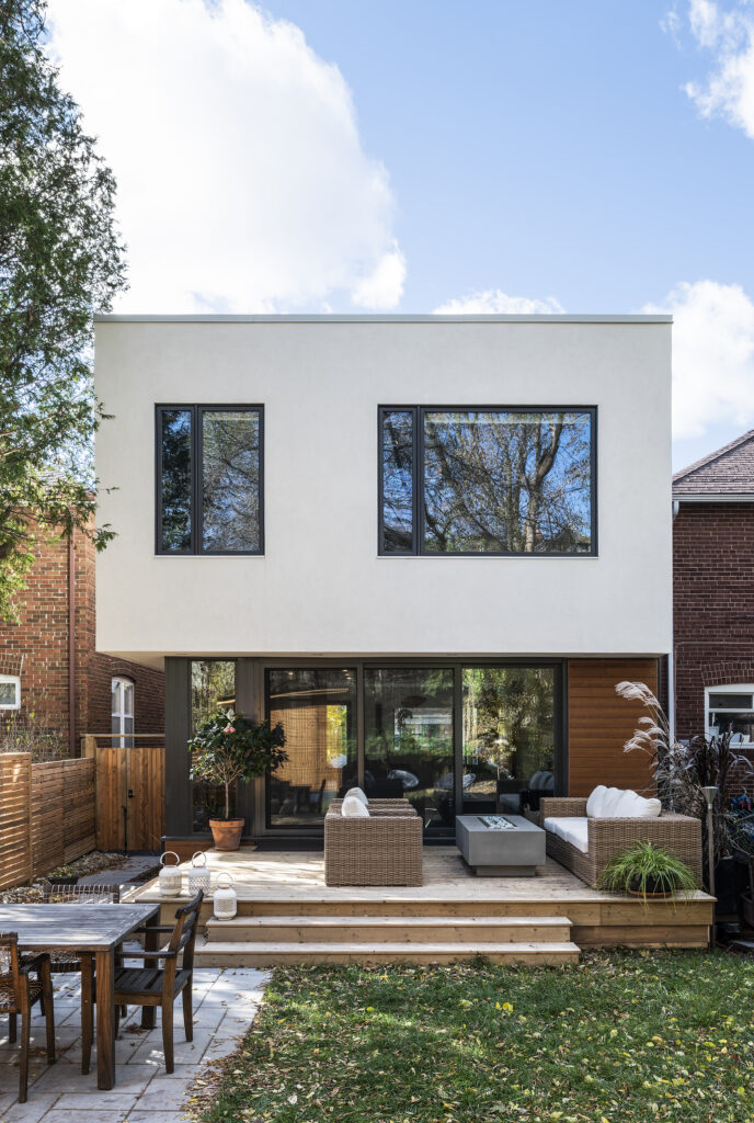 A modern two-story house with a white facade, large windows, and wooden accents features a spacious deck with outdoor seating. The backyard includes a dining area, green grass, and surrounding trees.