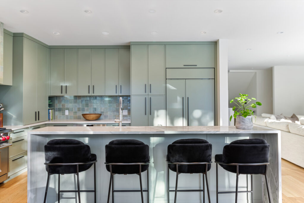 A modern kitchen with sage green cabinets, a marble island countertop, four black velvet barstools, a potted plant, and a tiled backsplash, with natural light streaming in from the right.