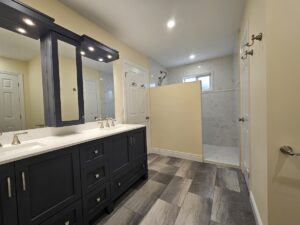 Modern bathroom with a double sink vanity, blue cabinets, large mirrors, and overhead lights. There is a walk-in shower with a half wall, light tile flooring, and beige walls. Towel hooks are mounted on the right wall.