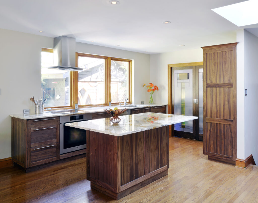 Modern kitchen with wooden cabinets, a marble island countertop, stainless steel appliances, hardwood floors, and large windows. A vase with orange flowers sits on the counter near decorative glass double doors.