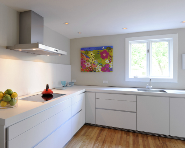 Modern kitchen with white cabinets, a stovetop with a red kettle, a bowl of fruit, a large window, and colorful floral artwork on the wall above the countertop. Light wood flooring.