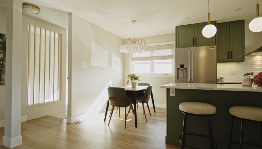 Bright modern kitchen and dining area with green cabinets, a round white light fixture, a window with blinds, a table with four chairs, two bar stools at an island, and light wood flooring.