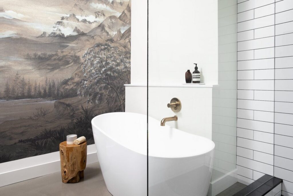 A modern bathroom with a white freestanding tub, brass fixtures, and white subway tile. A rustic wooden stool with a candle sits beside the tub. A mountain landscape mural decorates one wall.