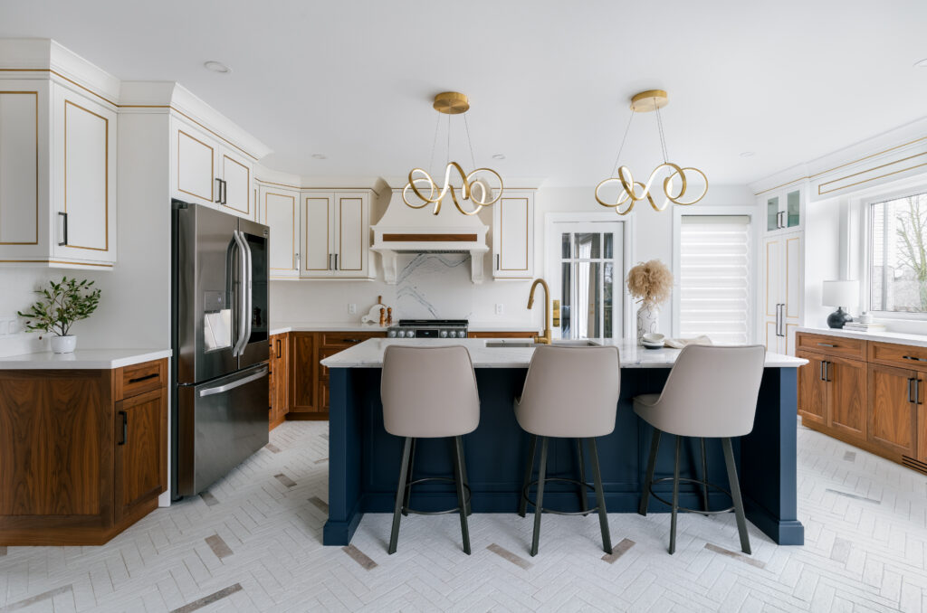 Modern kitchen with white and wood cabinetry, a navy island with three beige stools, gold light fixtures, stainless steel appliances, and a herringbone-patterned tile floor. Large windows provide natural light.
