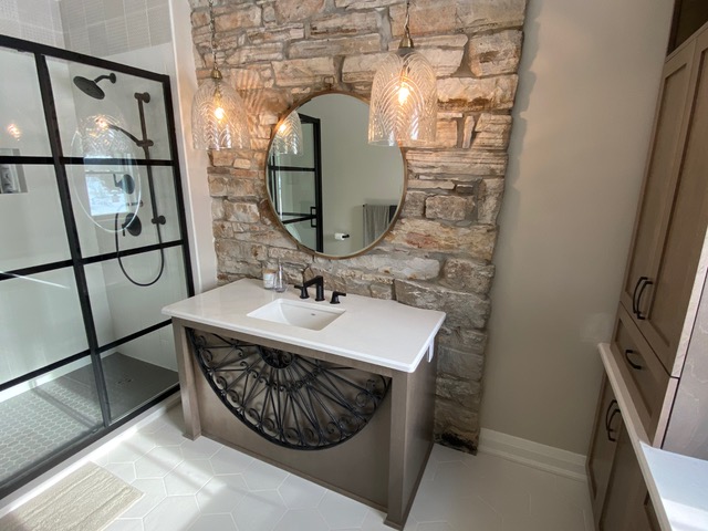 A bathroom features a large round mirror on a stone wall above a white sink with a black faucet. Two pendant lights hang from the ceiling, and a glass shower enclosure is visible on the left.