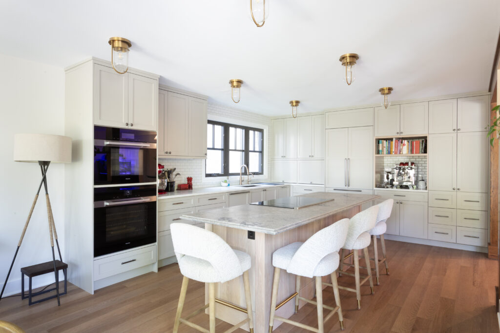 A modern kitchen with white cabinets, a large island with four cushioned stools, built-in ovens, pendant lights, and wooden floors. A floor lamp stands in the corner beside the oven.