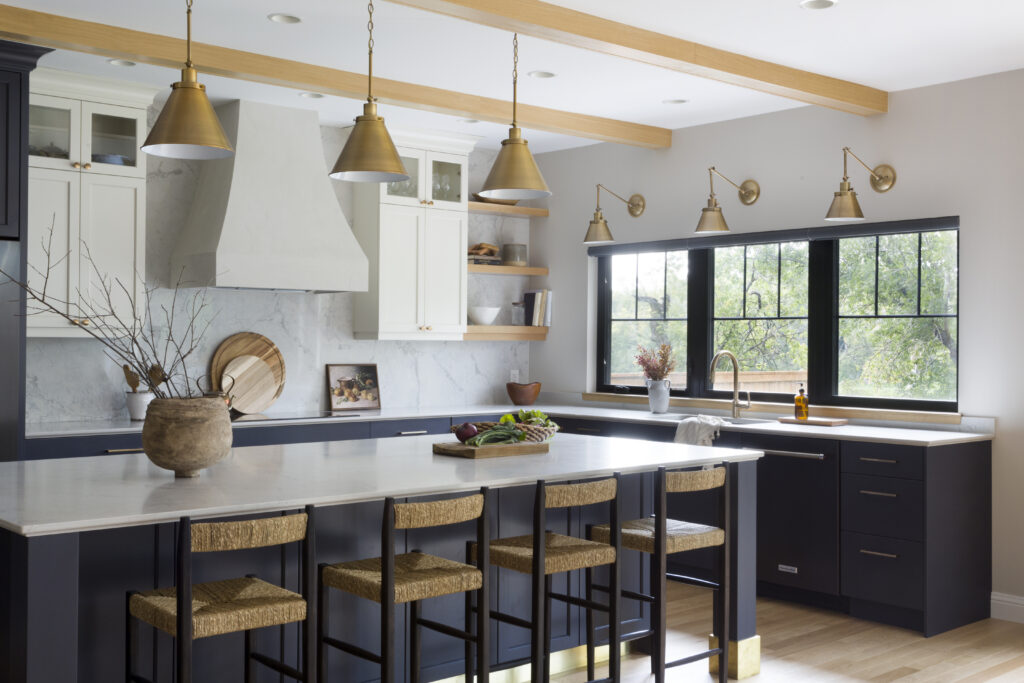 Modern kitchen with dark lower cabinets, white upper cabinets, a marble backsplash, a large island with woven barstools, gold pendant and wall lights, and large black-framed windows letting in natural light.