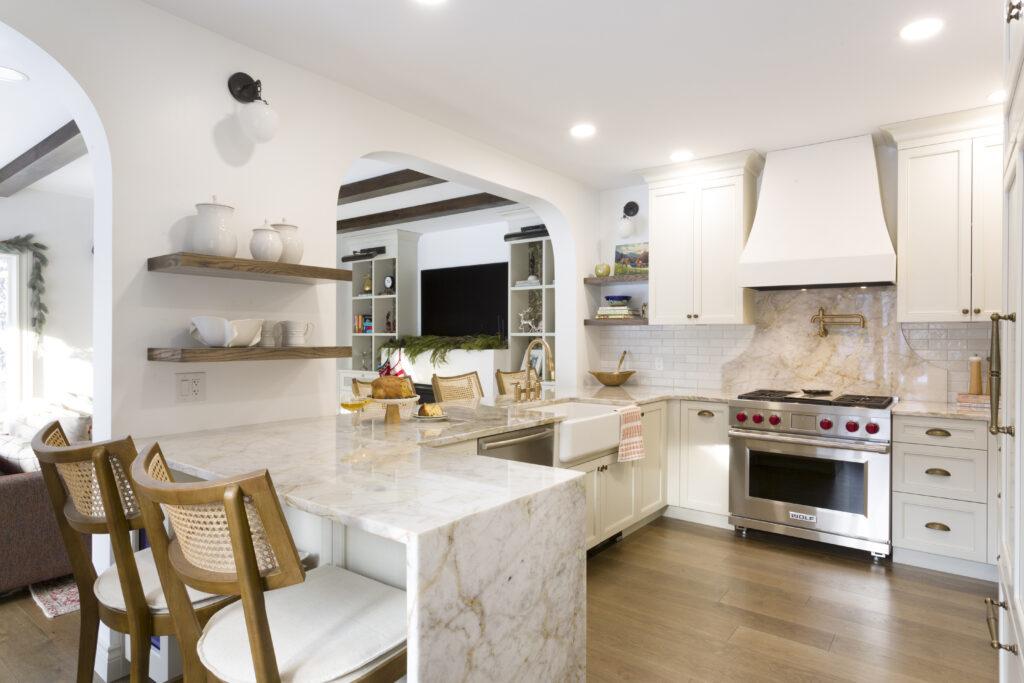 Bright, modern kitchen with white cabinets, marble countertops, open wooden shelves, and stainless steel appliances. Three wooden chairs line the island, and a cozy living area is visible through an archway.