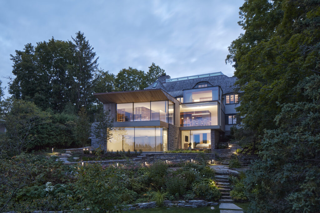 Modern multi-story house with large glass windows glowing with warm indoor lights at dusk, surrounded by lush greenery, stone steps, and a landscaped garden.