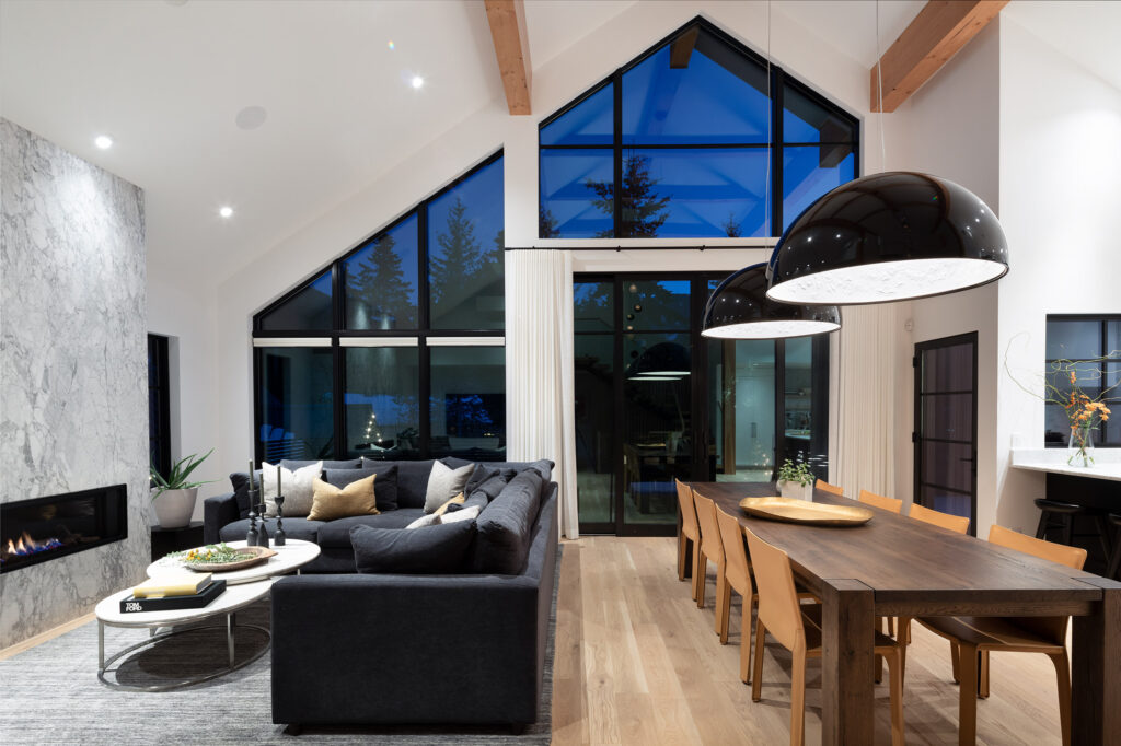 Modern open-concept living and dining area with vaulted ceilings, exposed wooden beams, large windows, a gray sectional sofa, marble fireplace, wooden dining table, and pendant lights. Evening sky is visible outside.