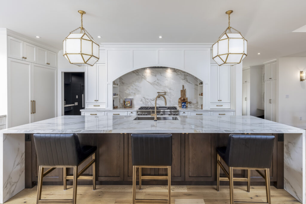 Modern kitchen with a large marble island, three black and gold bar stools, white cabinetry, a built-in stove, gold fixtures, and two geometric pendant lights hanging from the ceiling.