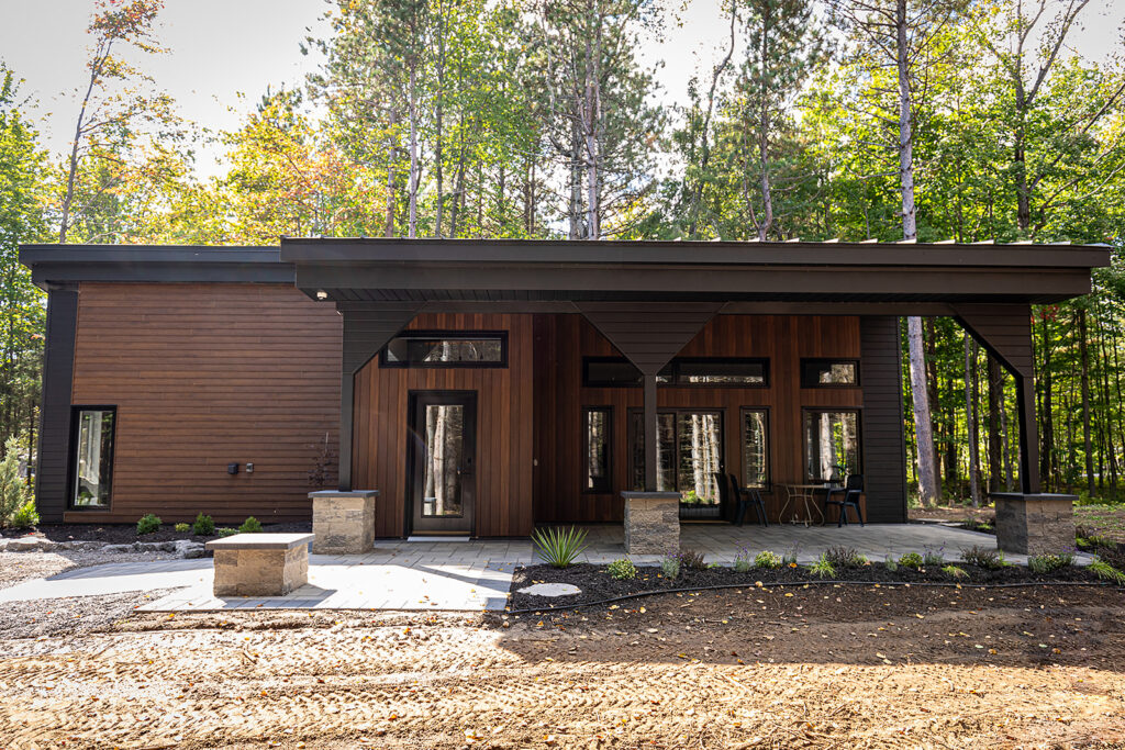 A modern, single-story house with dark wood siding and large windows, set in a forested area. The home features a slanted roof and a covered patio with stone pillars. Tall trees surround the property.