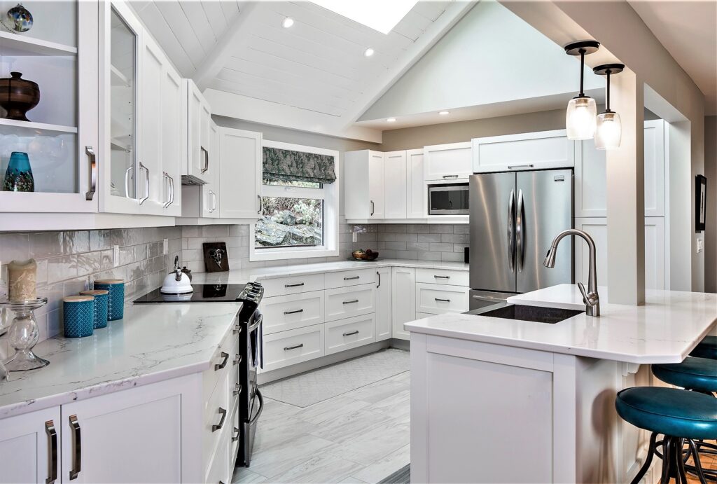 Modern kitchen with white cabinets, stainless steel appliances, marble countertops, pendant lights, bar with blue stools, and a window above the sink letting in natural light.