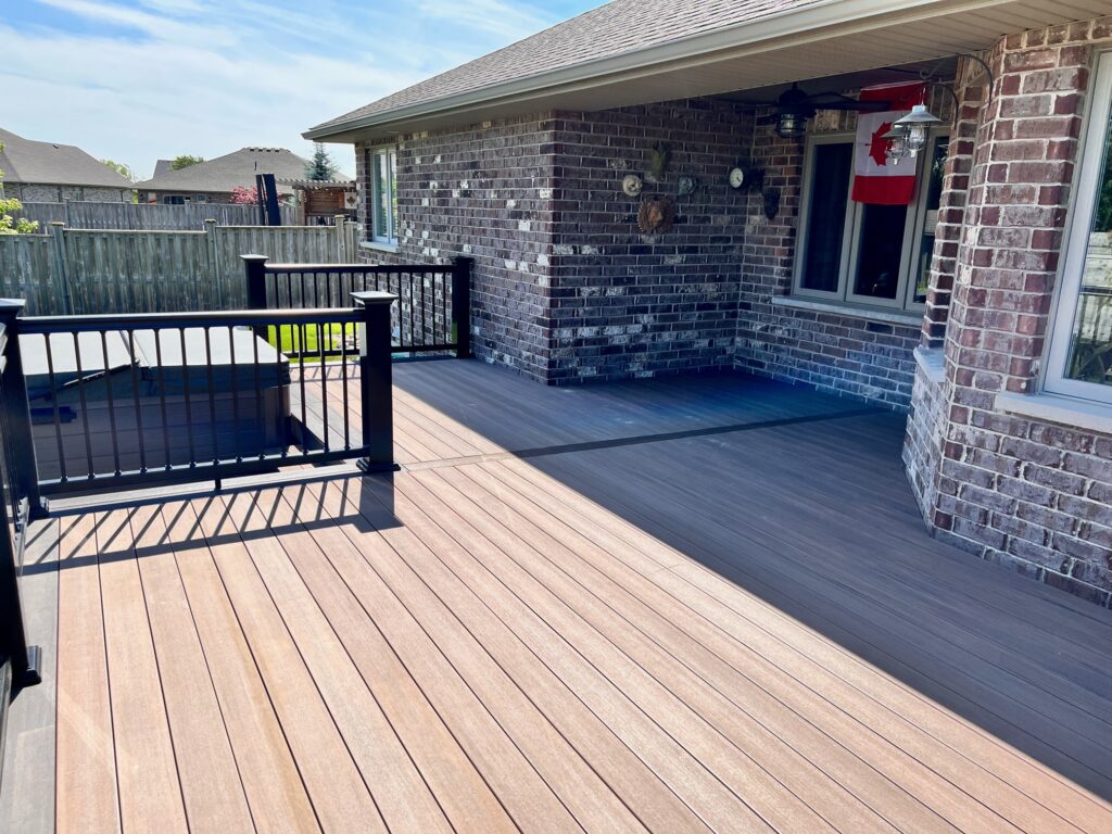 A spacious backyard deck with light brown wooden flooring, black railings, and attached to a brick house. There are two lanterns, a Canadian flag, and a hot tub visible on the deck under a sunny sky.