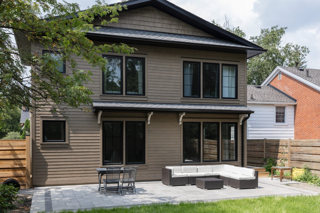 A modern two-story house with brown wooden siding, large black-framed windows, and a backyard patio featuring outdoor furniture including a sectional sofa, coffee table, and dining table with chairs. Trees and a wooden fence surround the yard.