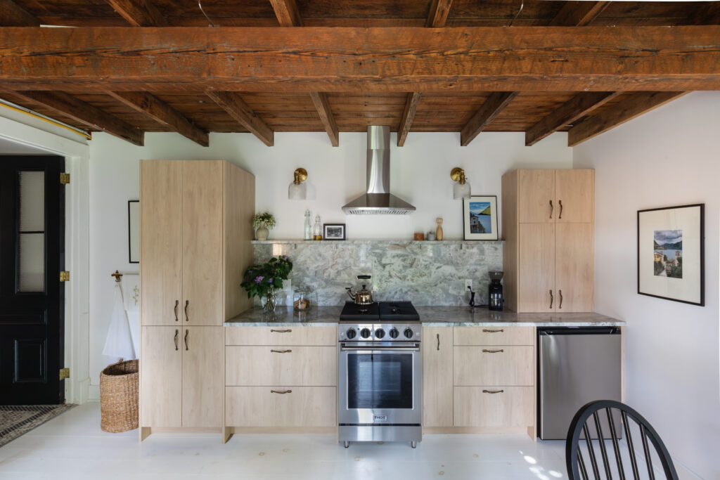 Modern kitchen with light wood cabinets, stainless steel oven and range hood, marble backsplash, mini fridge, and exposed wooden ceiling beams. Decor includes plants, wall art, and small kitchen appliances.