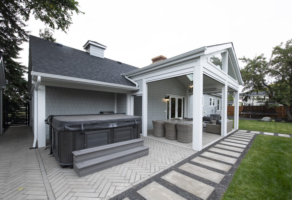 A modern backyard patio with a covered seating area, outdoor furniture, and a hot tub on a paved surface. Stepping stones lead across a grassy lawn beside the house.