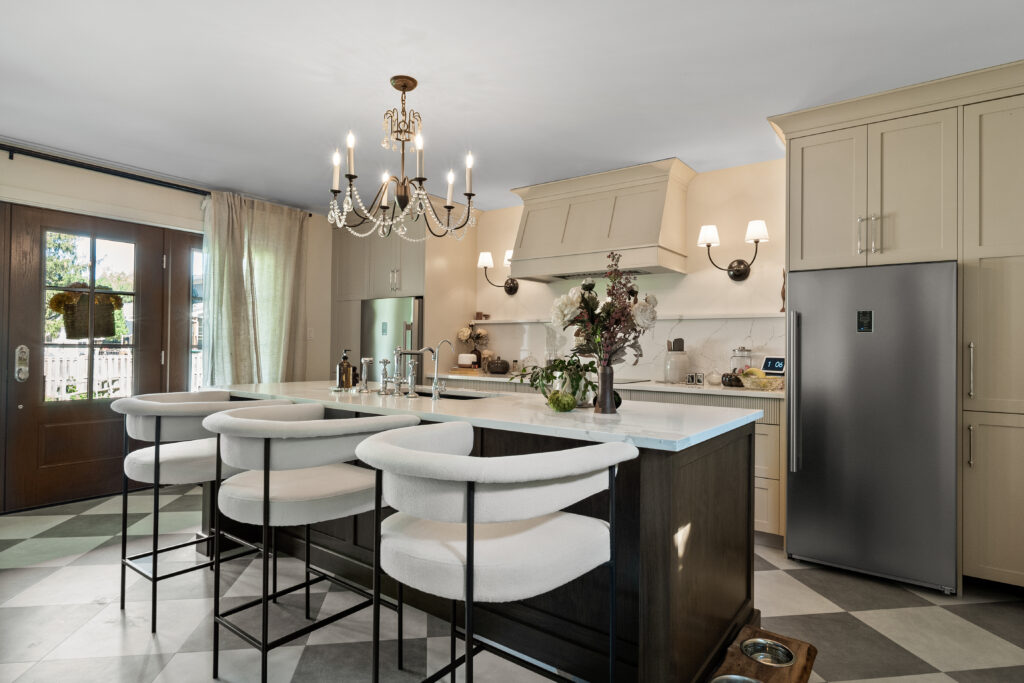 A modern kitchen with a large island, three cushioned bar stools, beige cabinets, stainless steel fridge, floral centerpiece, chandelier, and double glass doors letting in natural light.